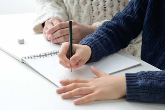 女性の隣でノートに文字を書きこむ男性の手元